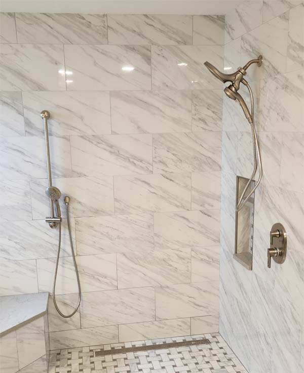 Newly installed brushed chrome shower fixtures in white marble stand up shower with dual shower heads and shower handles.